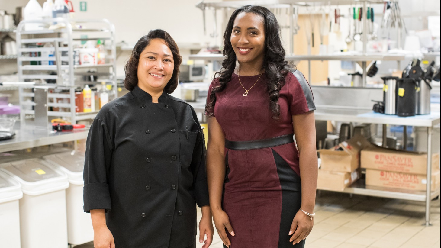 2 women (one wearing a black chef's jacket) in an industrial kitchen