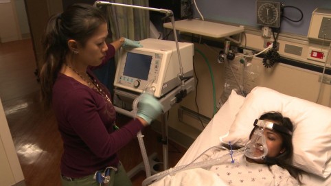 Health care worker standing next to a patient in a hospital bed wearing a breathing mask